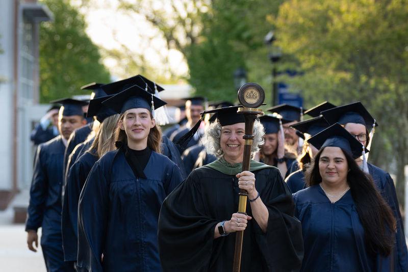 毕业生们头戴帽子，身穿长袍，排成两排在人行道上走着，由身穿黑袍的教员领着，手里拿着一根手杖和一个吊坠.